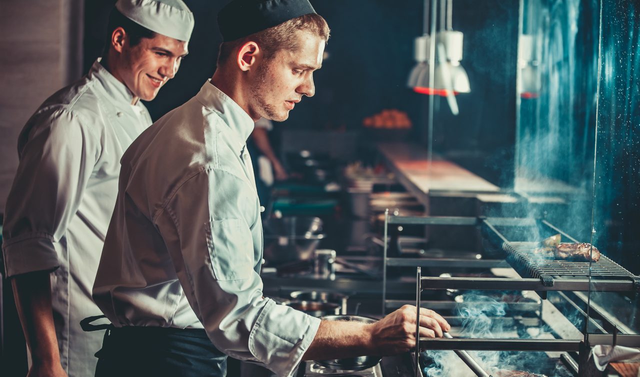 Food concept. Chef in white uniform monitors the degree of roasting and greases meat with oil in saucepan in interior of modern restaurant kitchen. Preparing traditional beef steak on barbecue oven.