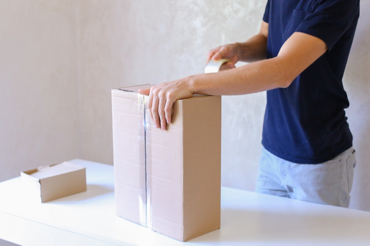 Deliveryman Standing in Room, Glue Tape Box and Takes Parcel in Hands and Smiling . Postman European Appearance in a Black T-Shirt and Cap Stands Near the Desk in the Office With White Walls and Unassisted Packaging Boxes, Adhesive Glue Tape on a Cardboard Box, the Courier Turns Box Vertically, Taking the Box in His Hands and Smiling at the Camera. Concept of Work in Office and Favorite Work 24 Hours Day, Mail Delivery From Post Office,free Delivery Worldwide or Delivery on Time, Advertising of New Shipping Service Company, Purchase at Any Time and Any Day.