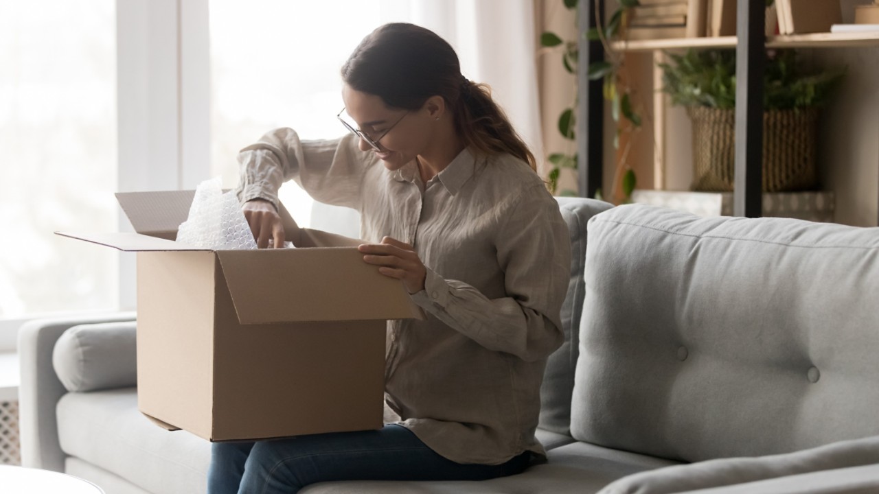 Excited young woman sit on couch in living room hold cardboard box unpacking delivered package, happy millennial girl in glasses open carton parcel at home unboxing goods shopping online